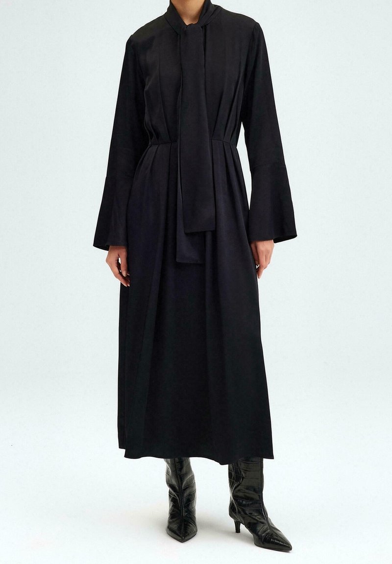 Femme portant une longue robe noire à manches évasées et nœud au cou, assortie de bottines à talons noires à bout pointu.