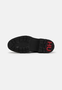 Semelle de chaussure noire avec motif de grip texturé et logo rouge "HU" sur la partie talon sur fond blanc uni.