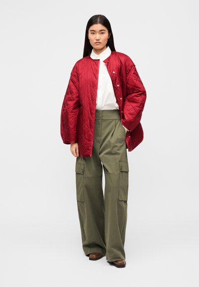 Κόκκινο υπερμεγέθες μπουφάν καπιτονέ με κλείσιμο με κουμπιά πάνω από λευκό πουκάμισο, συνδυασμένο με πράσινο λαδί cargo παντελόνι με φαρδιά πόδια, πλευρικές τσέπες και λεία υφή.