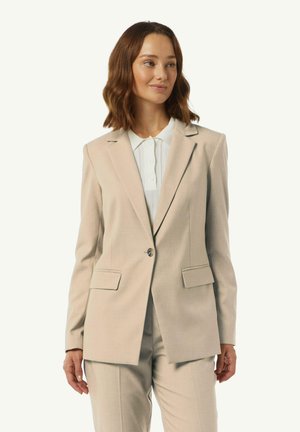 Vrouw met schouderlang golvend bruin haar draagt een beige tailored blazer en bijpassende broek over een wit overhemd met kraag, terwijl ze zijwaarts kijkt.