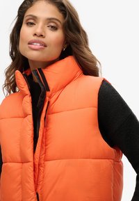 Heldere oranje puffer-vest met een hoge kraag, ritssluiting en zwarte accenten. Heeft een gestructureerd oppervlak met gewatteerde patronen.