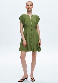 Grünes Leinenkleid mit kurzen Ärmeln, ausgestattet mit einem V-Ausschnitt, Knopfverschluss und Plissee-Design. Getragen mit schwarzen Sandalen.