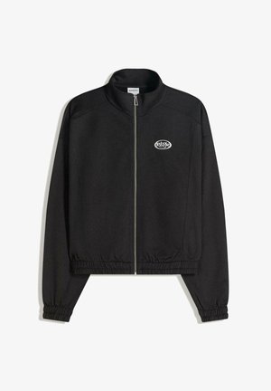 Veste noire zippée avec col montant, poignets et ourlet élastiqués, et un petit logo blanc sur le côté gauche de la poitrine.