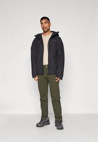 Muškarac u crnoj dukserici s kapuljačom, bež majici, maslinastozelenim hlačama i sivim planinarskim cipelama stoji naslonjen na jednostavnu bijelu pozadinu.