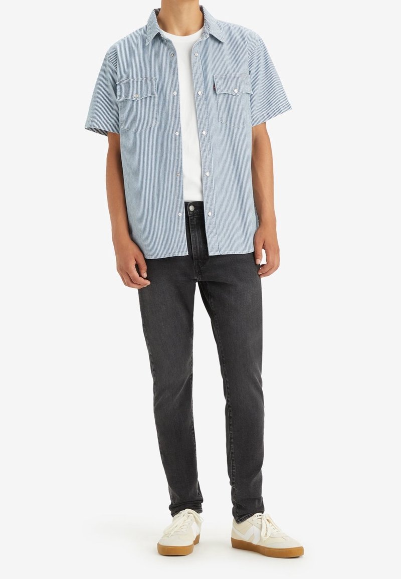 Homme portant une chemise à manches courtes rayée bleu clair boutonnée sur un t-shirt blanc, un jean noir et des baskets blanches à semelles en caoutchouc.