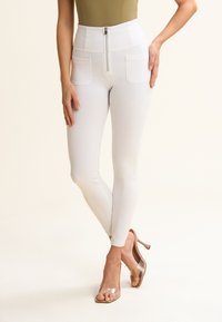 Leggings blancos de cintura alta con cremallera y dos bolsillos frontales, textura suave y silueta ajustada. Sandalias de tacón transparente.
