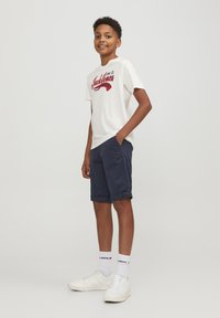 T-shirt chiara con testo grafico rosso, abbinata a pantaloni navy con risvolti, completata da sneaker bianche e calze.
