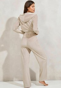 Béžová, mäkká súprava loungewear z tkaniny s kapucňou a širokými nohavicami, zobrazená zozadu s hladkou textúrou.