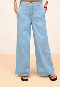 Pantalon large bleu clair en tissu denim, avec plis à l'avant, fermeture à boutons et poches latérales. Associé à des tongs noires.