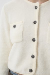 Krémově zbarvený vlněný kardigan s texturovanou tkaninou, dvěma předními kapsami a šedými knoflíky. Klasický design s kulatým výstřihem a zkráceným střihem.