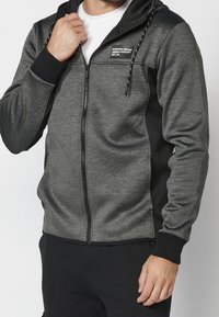 Sweat à capuche gris zippé avec des accents noirs, doté d'une capuche à cordon, de poches frontales et d'un tissu doux et texturé. Détail du logo sur la poitrine.