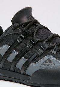 Czarne buty sportowe z teksturowaną cholewką, z trzema czarnymi paskami i szarym akcentem. Wytrzymała gumowa podeszwa i wyściełany kołnierz.