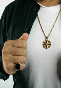 Collana in oro con pendente a forma di bussola dettagliato; anello ottagonale nero con accenti dorati; indossato sopra una maglietta bianca e una giacca nera.