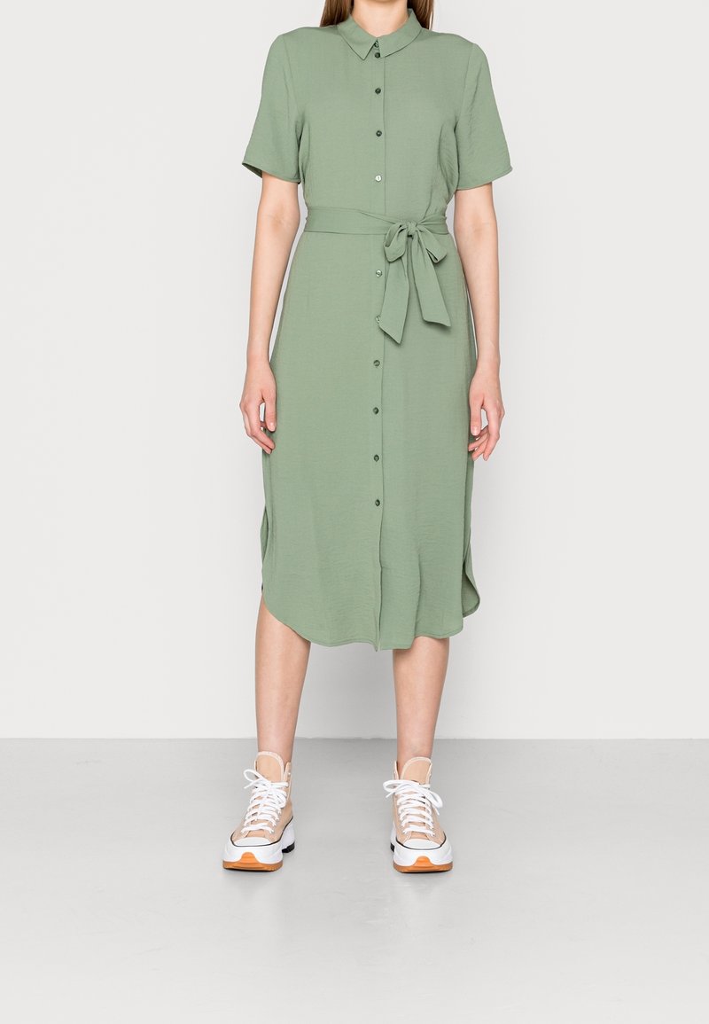 Grön, knälång klänning med korta ärmar, krage, knapplist framtill och knytliv. Bärs med vita och beige sneakers med orange detaljer.