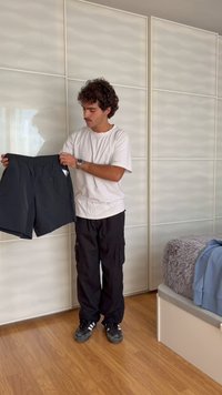 Άνδρας με λευκό μπλουζάκι και μαύρο παντελόνι κρατά μαύρα σορτς σε υπνοδωμάτιο με ξύλινο πάτωμα και κρεβάτι με γκρι κάλυμμα και μπλε φούτερ.