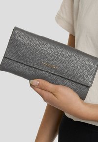 Cartera de cuero gris con textura de guijarros y cierre de solapa, con un sutil logo dorado. Forma compacta y rectangular con una superficie texturizada.
