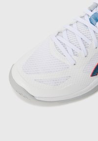 Baskets blanches avec une tige en mesh, présentant des accents bleus, une semelle en caoutchouc grise, et plusieurs œillets pour les lacets.