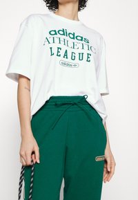 Valkoinen puuvillapaita, jossa on vihreä "adidas ATHLETIC LEAGUE" -teksti, yhdistettynä vihreisiin collegehousuihin, joissa on logopatch ja nauhavyötärö.