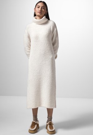 Robe pull blanche confortable à manches longues avec col montant, tissu texturé, coupe droite et fentes sur les côtés, associée à des chaussures marron à enfiler.