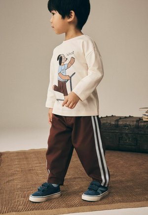 Jong kind draagt een beige shirt met lange mouwen met een hond op een scooter en een bruine broek met witte strepen, staat op een gestructureerd vloerkleed binnen.