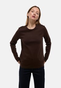 Chemise marron à manches longues avec un col rond, fabriquée dans un tissu lisse et légèrement extensible. Portée sur un jean foncé, avec une coupe décontractée.