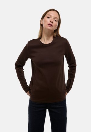 Chemise marron à manches longues avec un col rond, fabriquée dans un tissu lisse et légèrement extensible. Portée sur un jean foncé, avec une coupe décontractée.