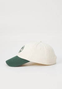 Cremefarbene Baseballmütze mit einem dunkelgrünen Schirm, ausgestattet mit einem gestickten runden Logo auf der Vorderseite und Belüftungslöchern oben.