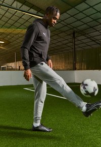 Zwarte hoodie en grijze joggingbroek gedragen door een persoon die binnen op kunstgras soccer speelt, terwijl hij een meerkleurige voetbal trapt.