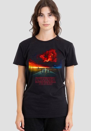 Woman wearing a black t-shirt featuring four kids on bikes on a road under a fiery red and orange sky with "Stranger Things" text below.