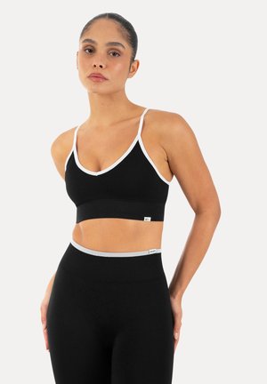 Kvinde iført sort sports-bh og højtaljede leggings med hvide kanter, stående mod en ensfarvet lys baggrund.