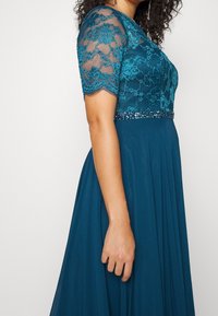 Femme en robe bleu sarcelle avec manches courtes en dentelle, broderie florale et une ceinture perlée, debout devant un fond uni.