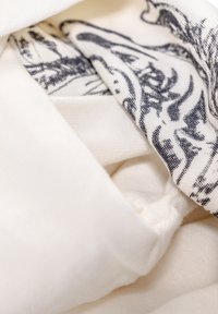 Tessuto bianco con una finitura testurizzata, sovrapposto a una sciarpa bianco e nera con motivi astratti. Aspetto morbido e drappeggiato.