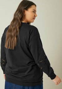 Svart sweatshirt med broderade utskärningar på ärmarna, avslappnad passform och ribbade muddar. Tillverkad av ett mjukt material med en slät yta.