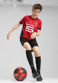 Raudona ir juoda futbolo apranga su raštuotu marškinėliu ir logotipu, juodos šortai, juostos iki kelių juodos kojinės ir juodi futbolo batai, šalia raudono ir juodo futbolo kamuolio.