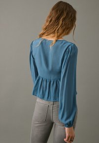 Blusa teal con maniche lunghe e arricciate e vita a peplo, realizzata in tessuto leggero. Presenta un ampio scollo e una superficie testurizzata.