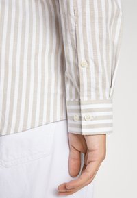 Beige and white striped shirt sleeve with two button closure, featuring a smooth texture and contrasting stripes at the cuff.