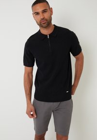 Sort polo-shirt med korte ærmer, tekstureret mønster, lynlås-halskrave og ribbet kant. Modellen har grå shorts på og viser en afslappet stil.