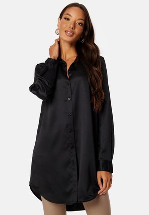 Zwarte satijnen blouse met lange mouwen, een puntige kraag en een gebogen zoom. Heeft een gladde textuur en subtiele glans.