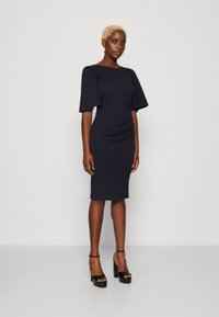 Jūras zilas, pielāgotas kleitas ar īsām pūkainām piedurkn�ēm, teksturētu audumu, sānu ručinga detaļu un ceļa garuma griezumu, apvienotas ar melniem platformu papēžiem.