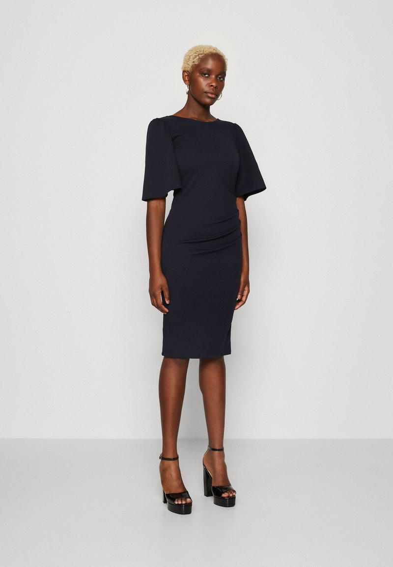 Jūras zilas, pielāgotas kleitas ar īsām pūkainām piedurknēm, teksturētu audumu, sānu ručinga detaļu un ceļa garuma griezumu, apvienotas ar melniem platformu papēžiem.