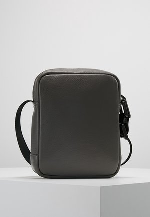 Kleine donkergrijze leren crossbodytas met een zwarte verstelbare riem en ritssluiting, geplaatst op een wit voetstuk tegen een effen achtergrond.