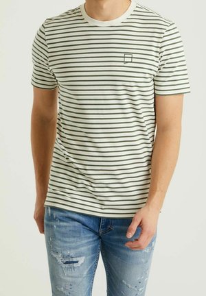 Homme portant un t-shirt blanc à rayures horizontales vertes et un petit logo sur la poitrine, associé à un jean bleu clair déchiré, debout devant un fond uni.