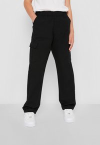 Pantalons cargo noirs avec une taille élastique, poches latérales et une coupe décontractée, associés à des baskets blanches sur un fond neutre.