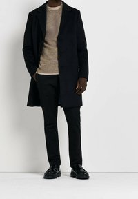 Homme portant un manteau noir, un pull beige, un pantalon noir et des mocassins noirs, debout devant un fond blanc avec une main dans la poche.
