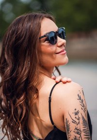 Žena s dlouhými hnědými vlasy a květinovými tetováními na rameni nosí černé sluneční brýle a černé šaty na ramínka, usmívá se a dívá se do strany.