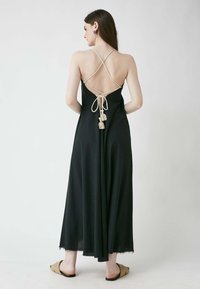 Schwarzes Maxikleid mit einem rückenfreien Design, ausgestattet mit überkreuzten Trägern und Quastenakzenten, aus weichem Stoff gefertigt, am Saum mit fransigem Rand.