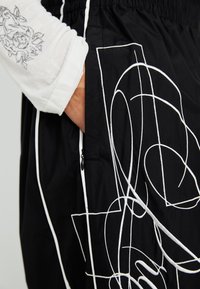 Black pant featuring white abstract line designs, elastic waistband, side pocket with zipper, and contrasting stitching detail.