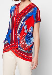 Blouse rouge à manches courtes avec des motifs floraux et géométriques bleus. Présente un décolleté en V, un design fluide et des rayures contrastées bleu marine et blanches.