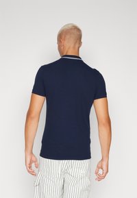 Námořnická modrá polokošile s jemnou strukturou, krátkými rukávy a klasickým límečkem s bílými akcenty. Nosená s pruhovanými kalhotami.