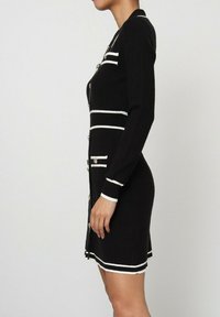 Robe en tricot noire avec des manches longues, des rayures horizontales blanches, des boutons argentés, et une silhouette ajustée avec un motif texturé.